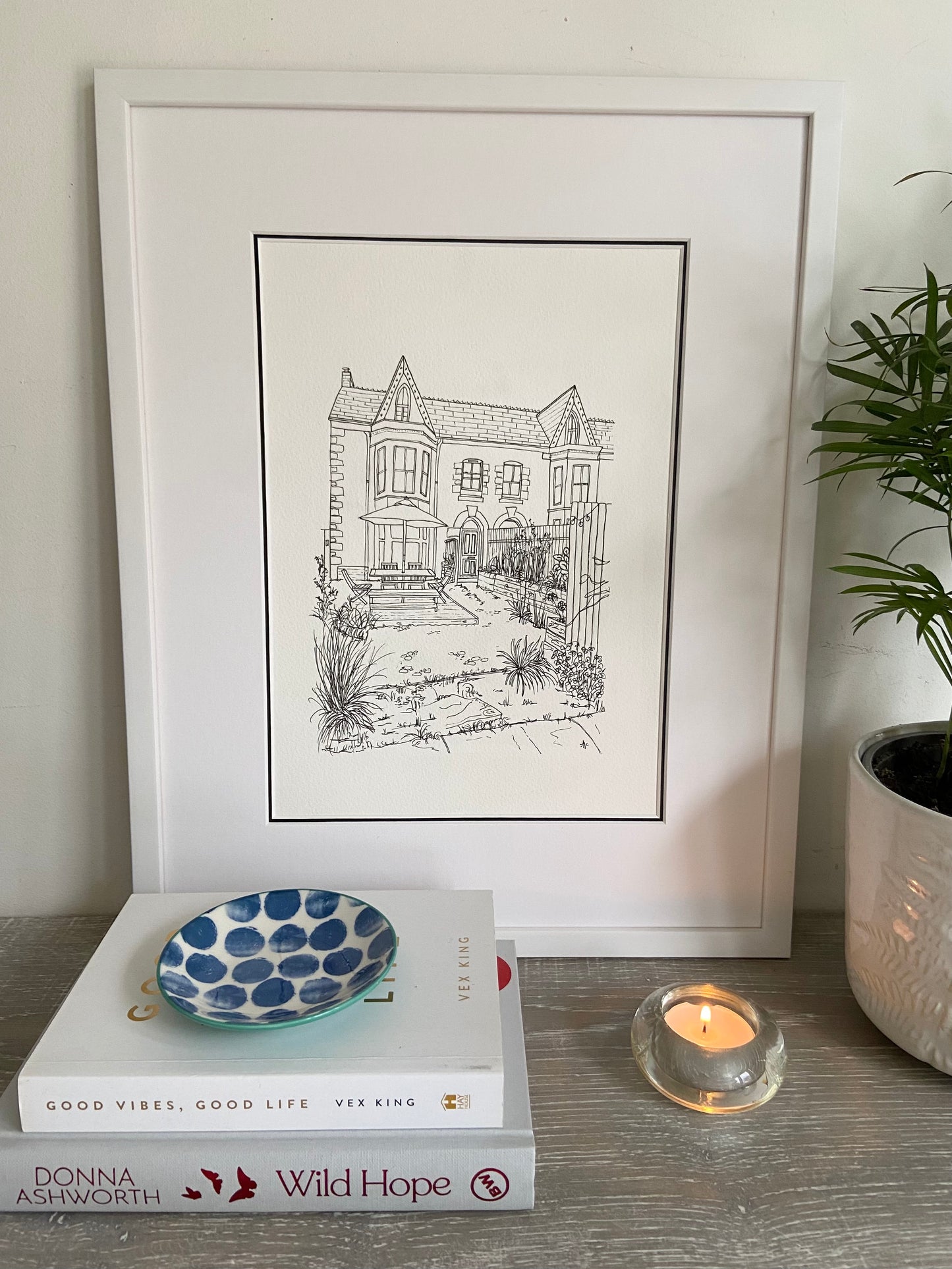 A framed hand-drawn illustration of a house, displayed on a shelf with books, a bowl, and a candle, next to a potted plant.