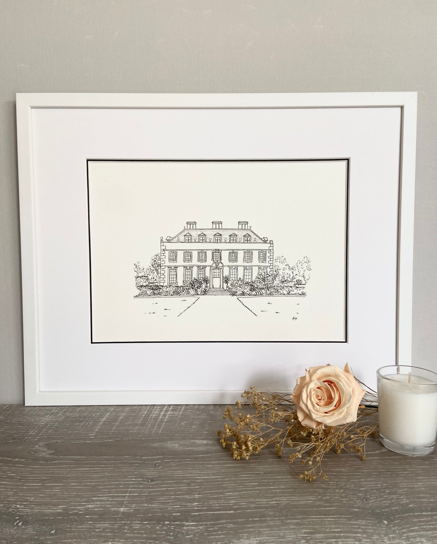 A framed hand-drawn line illustration of a wedding venue, displayed on a table with a white rose and a candle beside it.