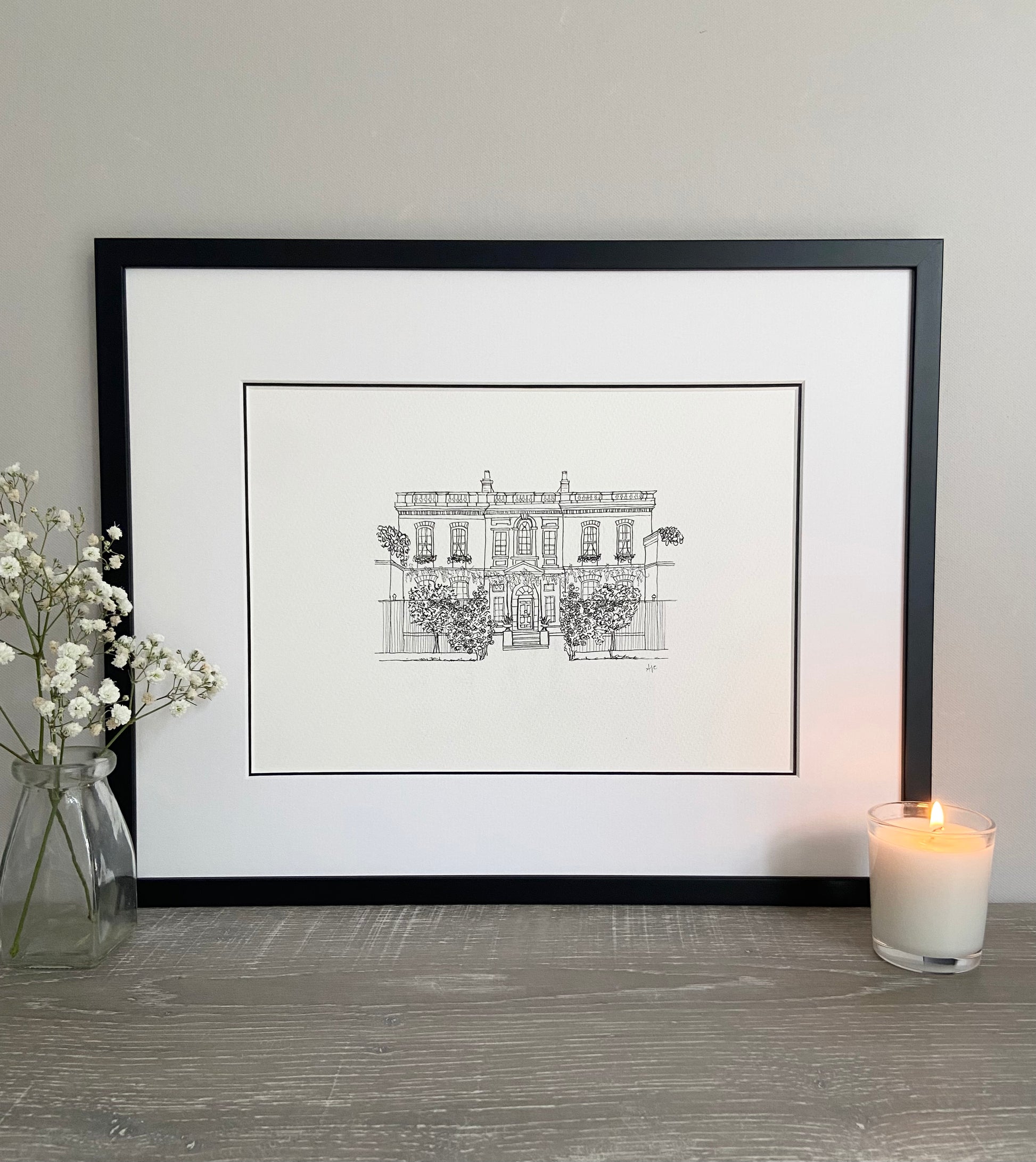 Bridgerton House print in a black frame and double mounted, leaning against a wall with a lit candle and a vase of flowers either side.