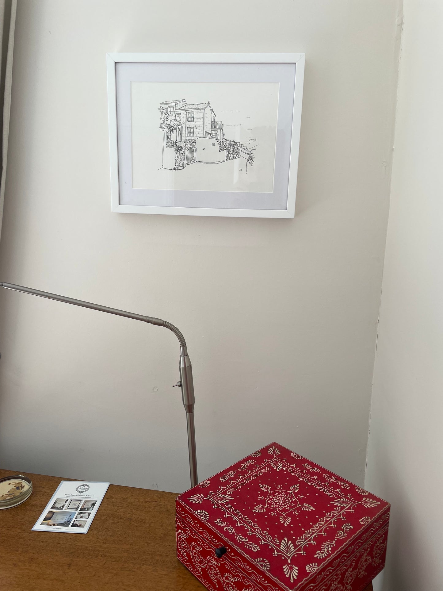 Framed black and white drawing on a wall above a red box and lamp.