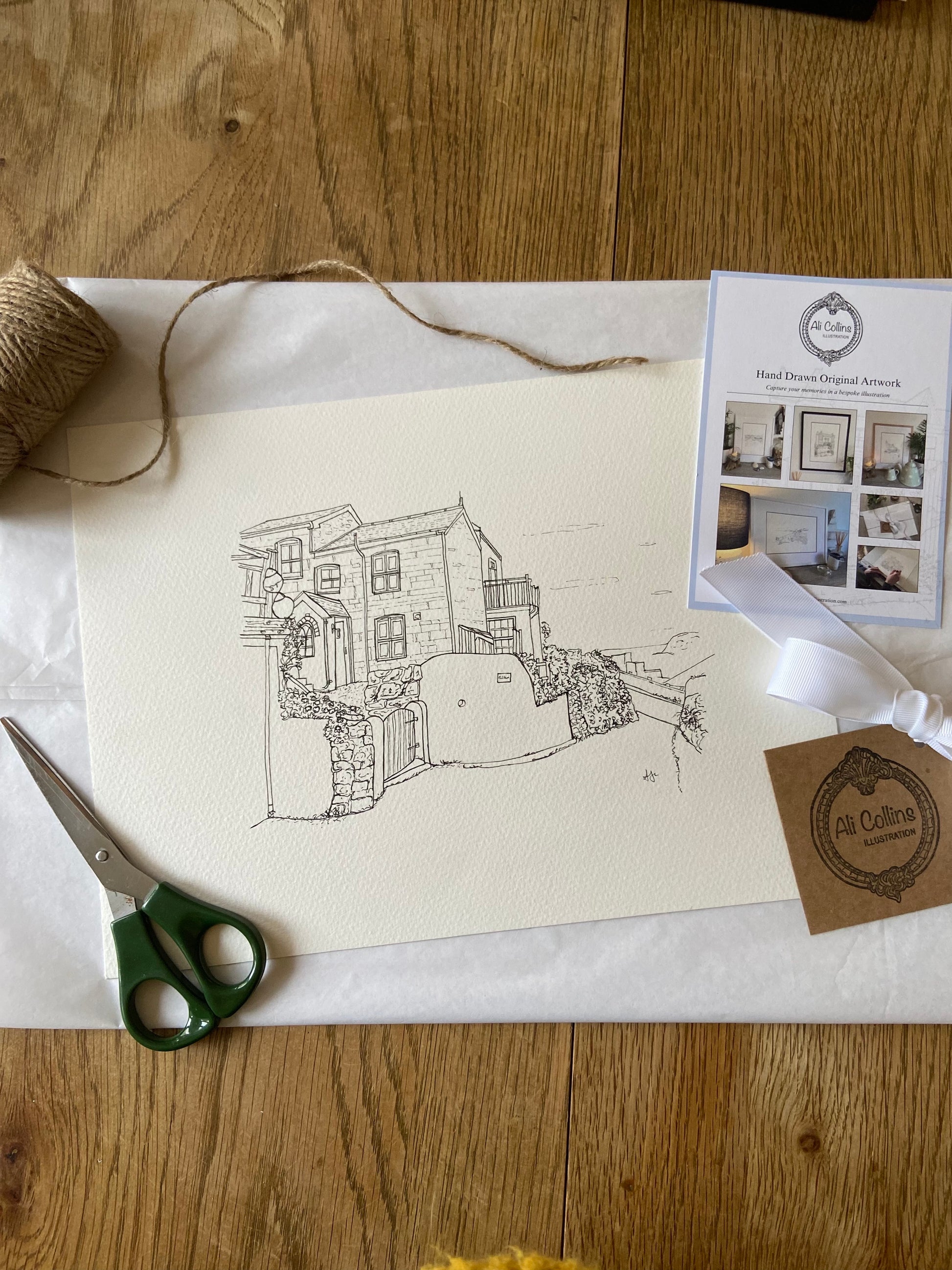 Artistic drawing of a house on paper with scissors, twine, and a brand card on a wooden surface.