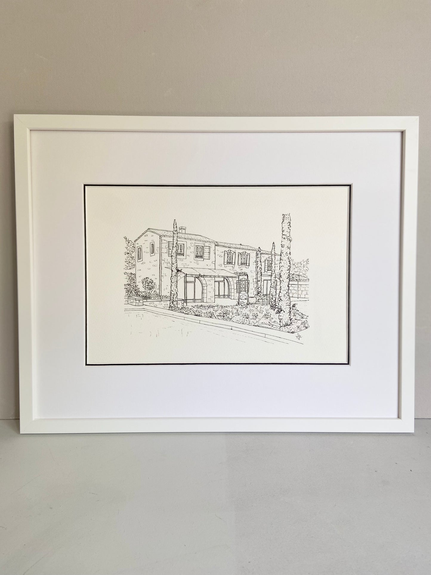 Framed black and white line drawing of a house on a plain wall.