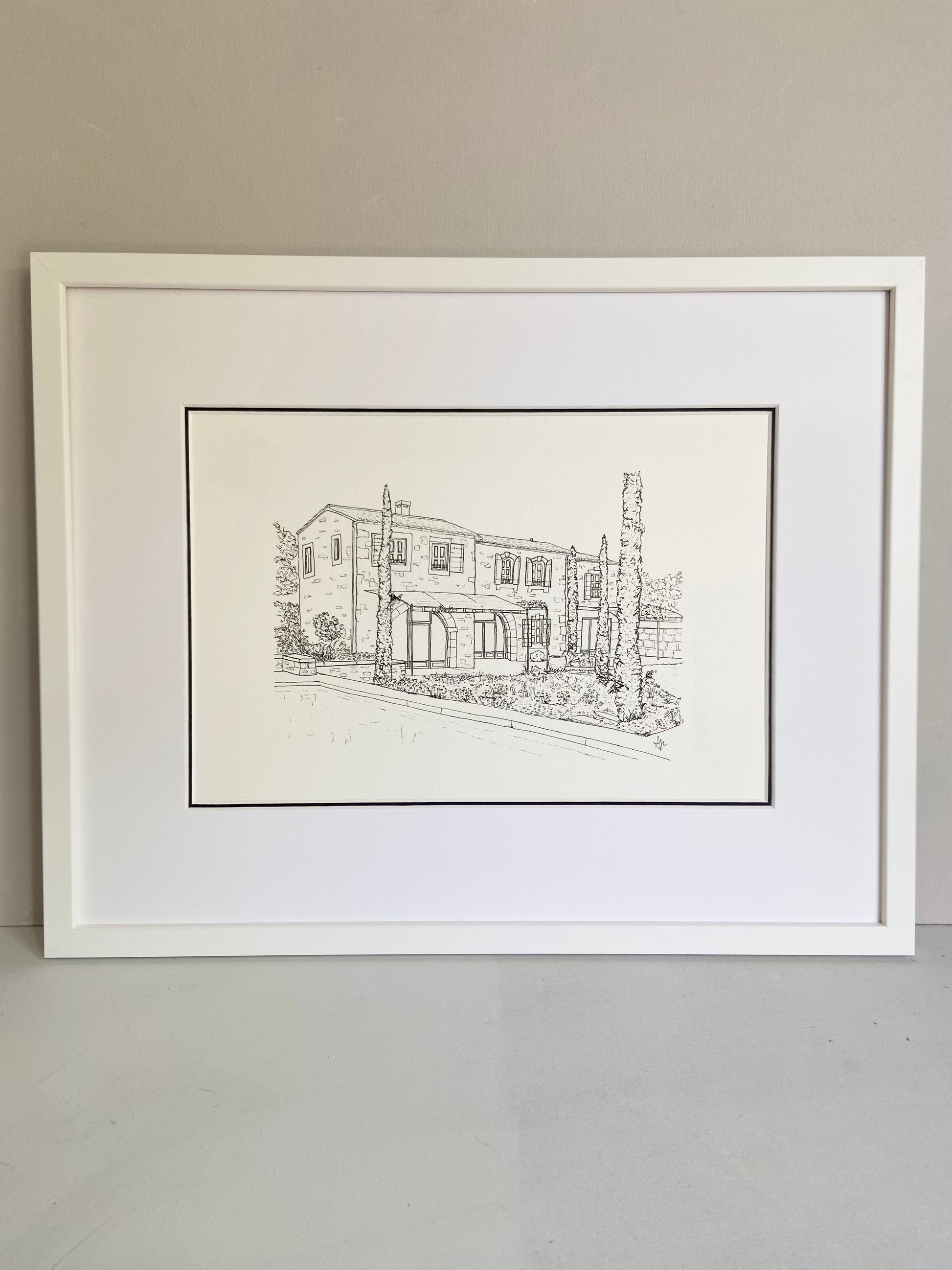 Framed black and white line drawing of a house on a plain wall.
