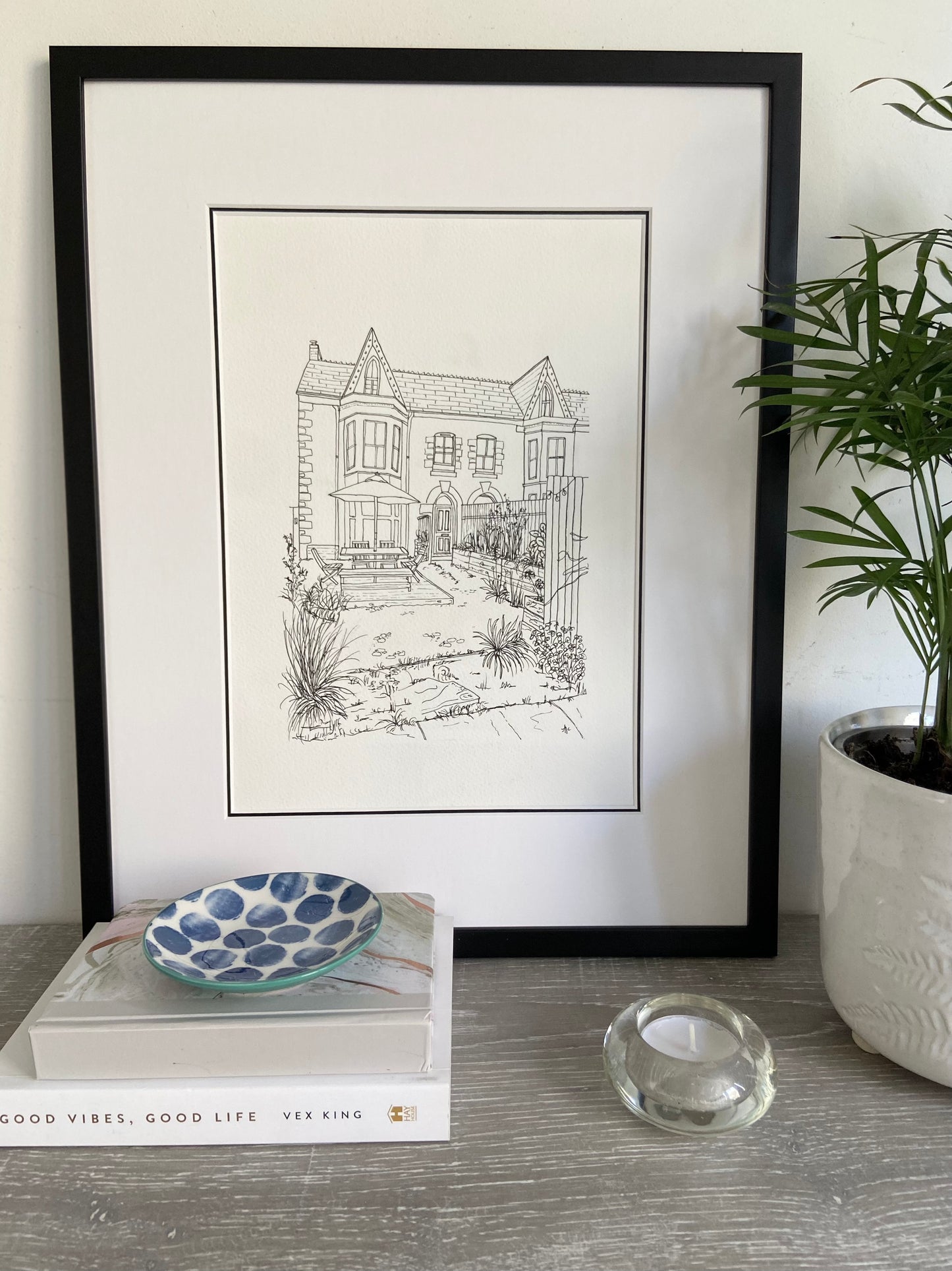 A framed hand-drawn illustration of a house, displayed on a shelf with books, a bowl, and a candle, next to a potted plant.