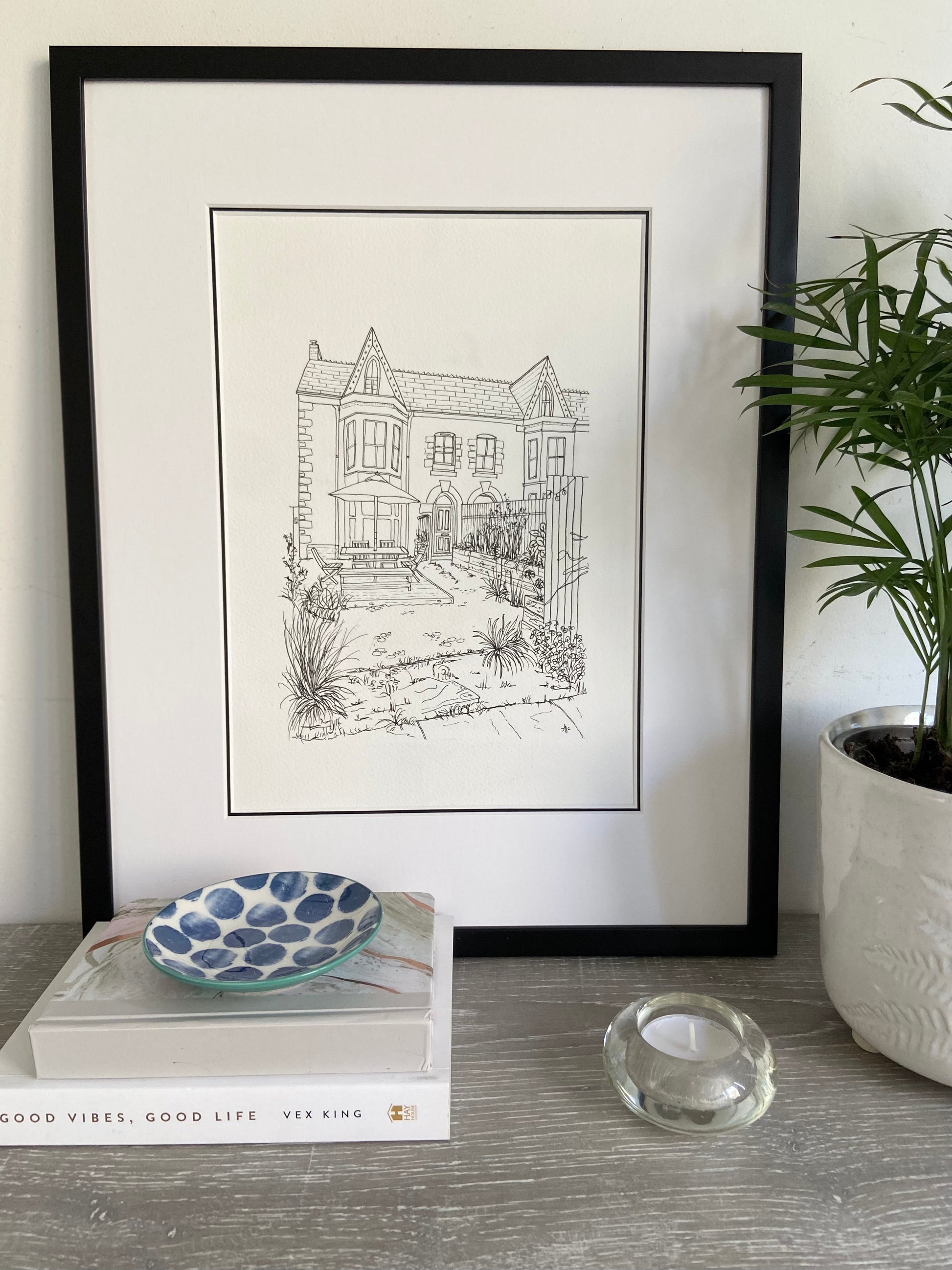 A framed hand-drawn illustration of a house, displayed on a shelf with books, a bowl, and a candle, next to a potted plant.