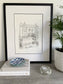 A framed hand-drawn illustration of a house, displayed on a shelf with books, a bowl, and a candle, next to a potted plant.
